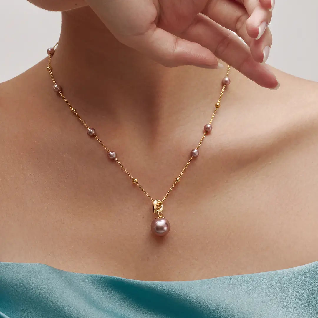 A close-up of a model's neck and upper chest wearing a delicate gold chain necklace with small pinkish-purple pearls spaced along the chain and a larger matching pearl pendant at the center.
