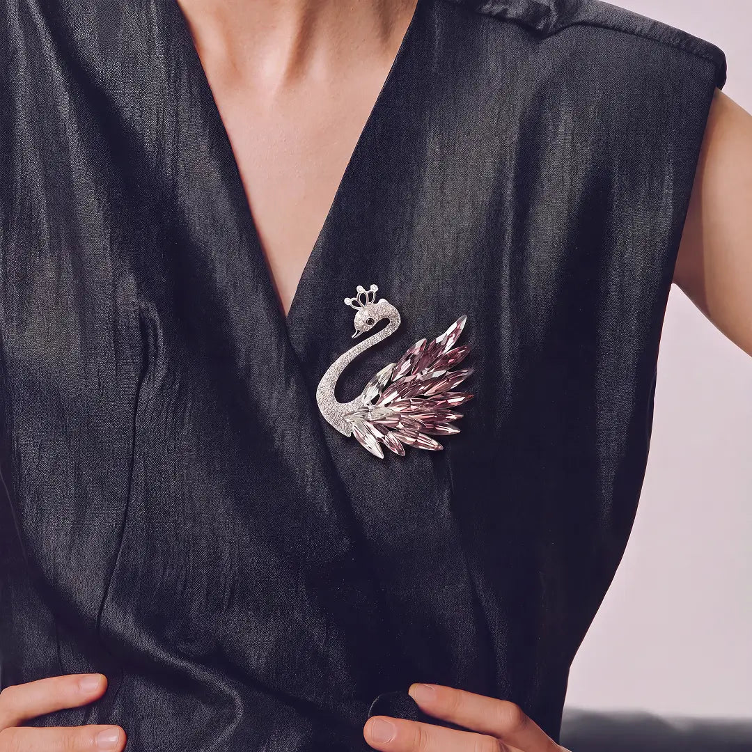 A silver-toned peacock brooch with faceted pink and purple crystals, pinned to the chest of a black satin V-neck dress.
