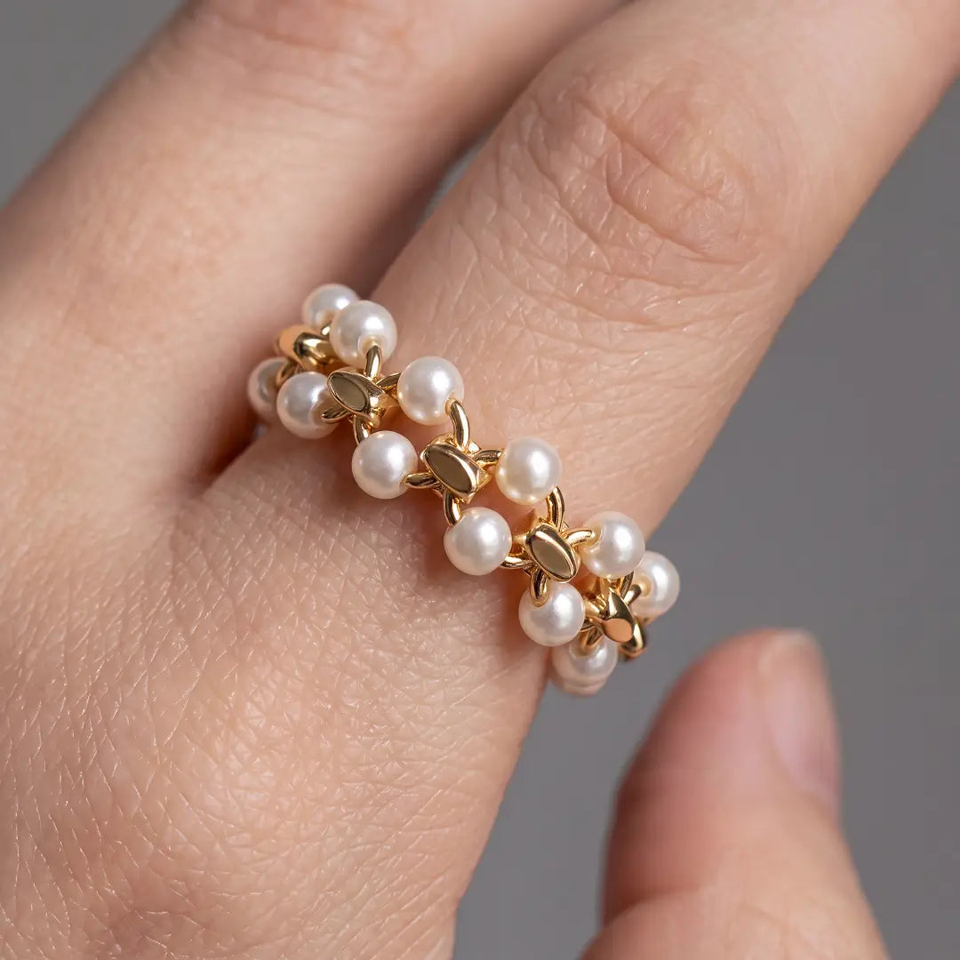 A close-up of a hand wearing a delicate gold ring adorned with small white pearls and tiny gold floral details, set against a soft gray background.