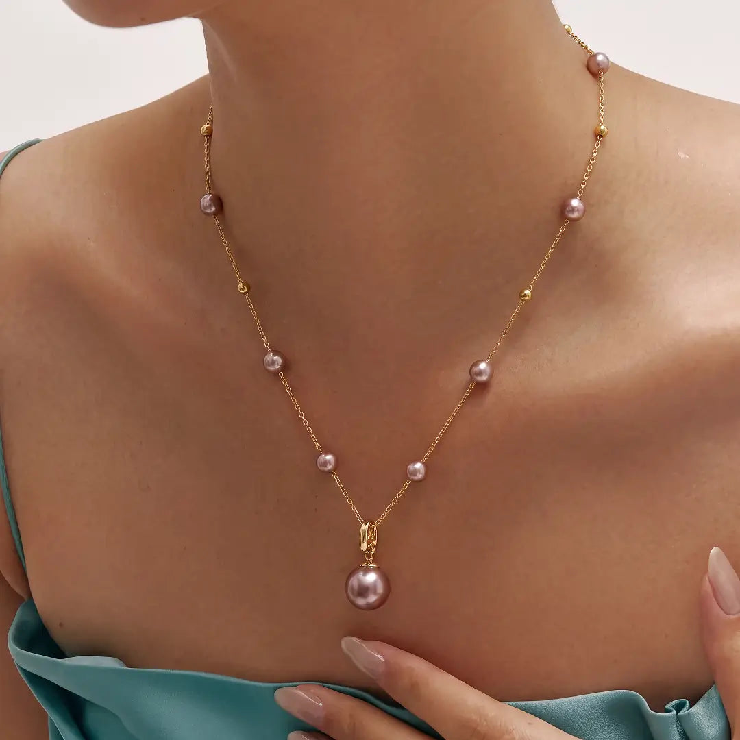 A gold chain necklace with scattered pink freshwater pearls and a larger pendant pearl, shown worn on a model's neck.