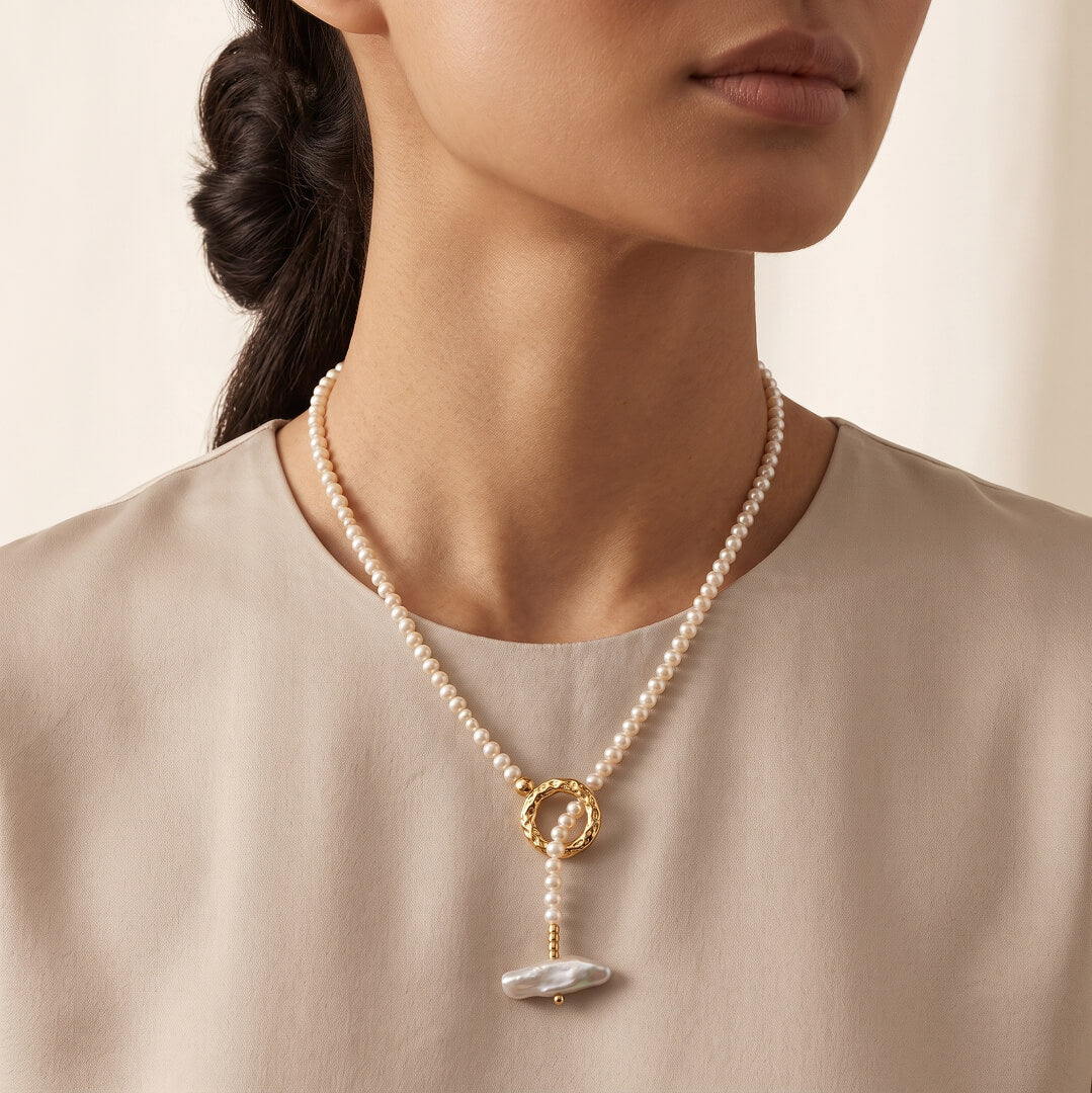 A close-up of a model wearing a delicate pearl necklace with a gold ring clasp and a white mother-of-pearl pendant, paired with a beige top.
