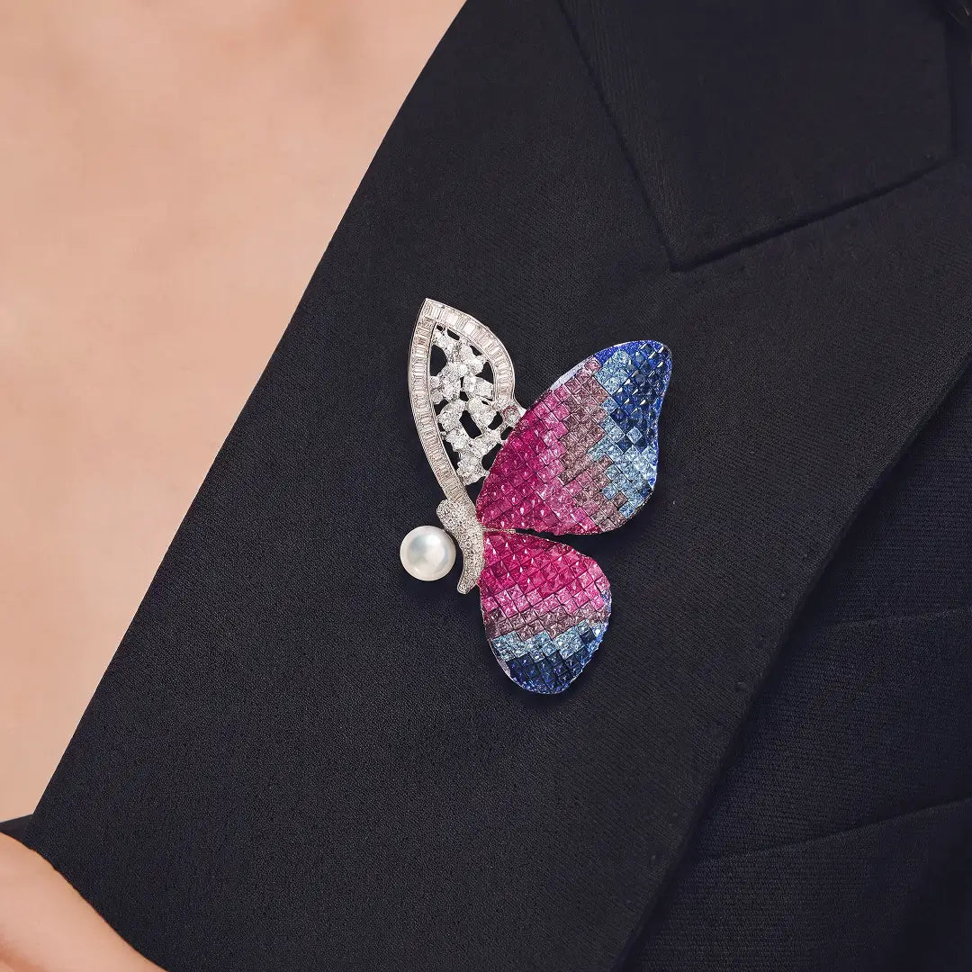 A butterfly-shaped brooch with textured pink and blue wings, a silver filigree back, and a single pearl accent, pinned to a black blazer.