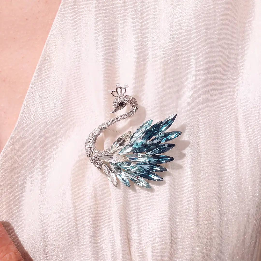 A swan-shaped brooch with silver-toned metal and faceted blue and clear crystals, displayed on a soft white draped fabric.