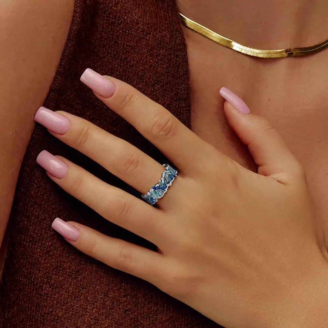 A close-up of a hand with pink manicured nails wearing a silver ring set with blue marquise-cut crystals, styled with a gold necklace against a brown knit top.