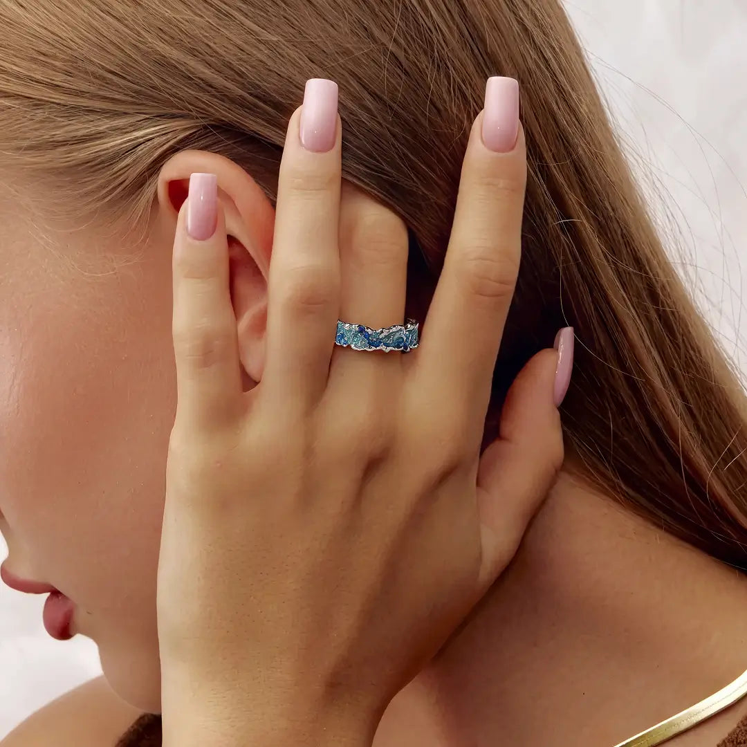 A close-up lifestyle shot of a woman's hand with pink manicured nails wearing a textured silver band ring set with light blue stones, against her ear and brown hair.