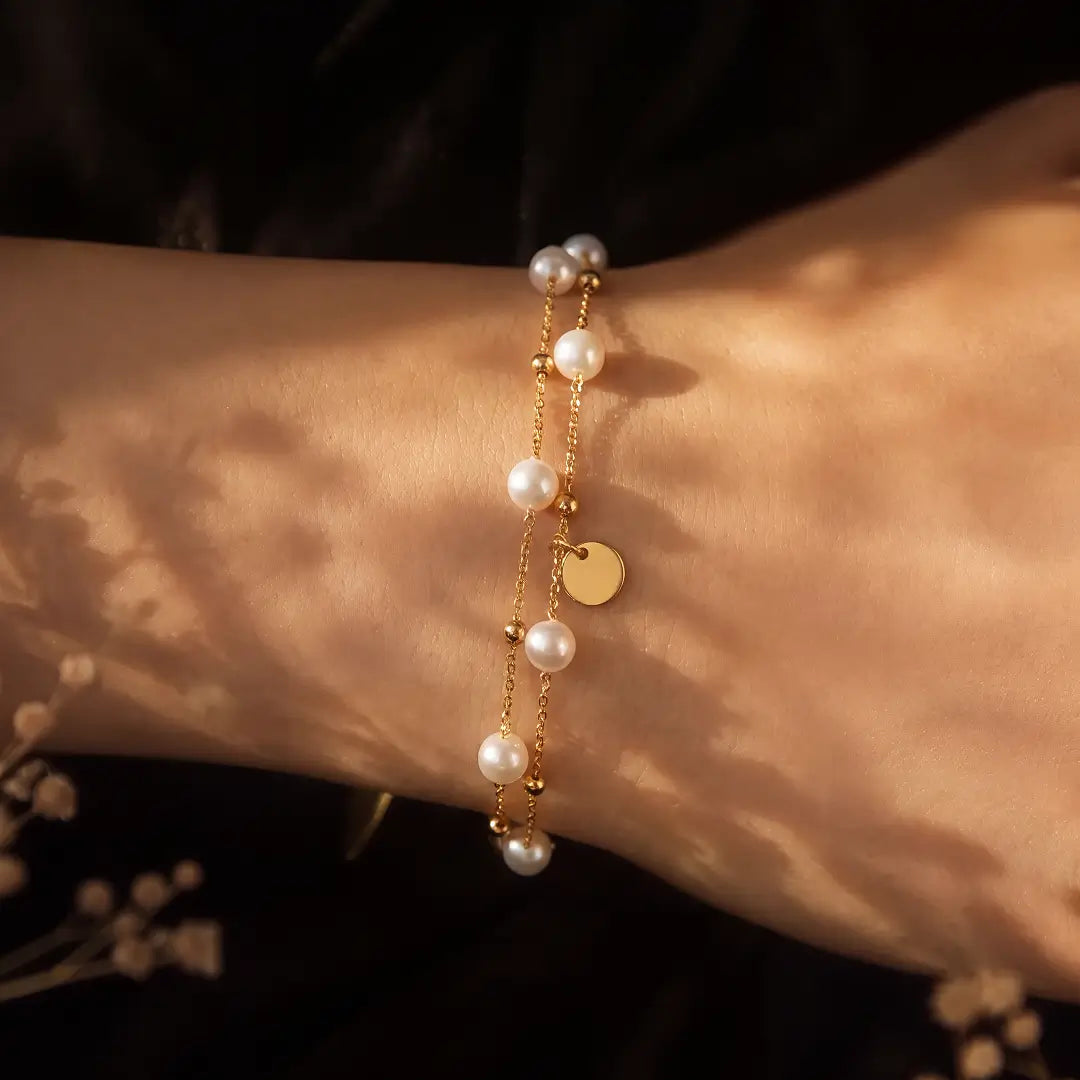 A delicate gold chain bracelet with white faux pearls and a small round gold disc charm, shown worn on a person's wrist under warm, dappled sunlight.