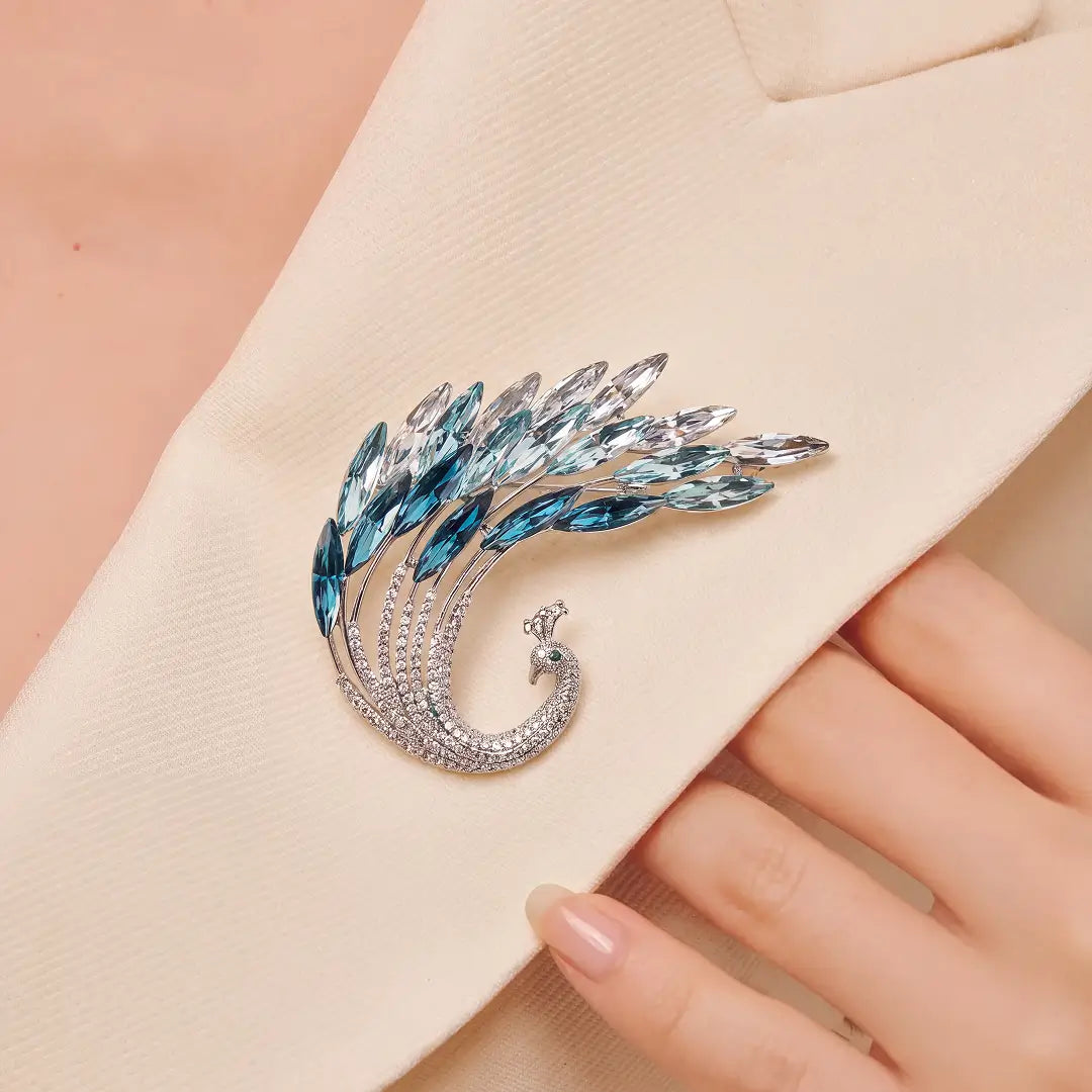 A silver-toned peacock feather brooch with blue and clear rhinestones, pinned to a beige blazer and held by a hand.