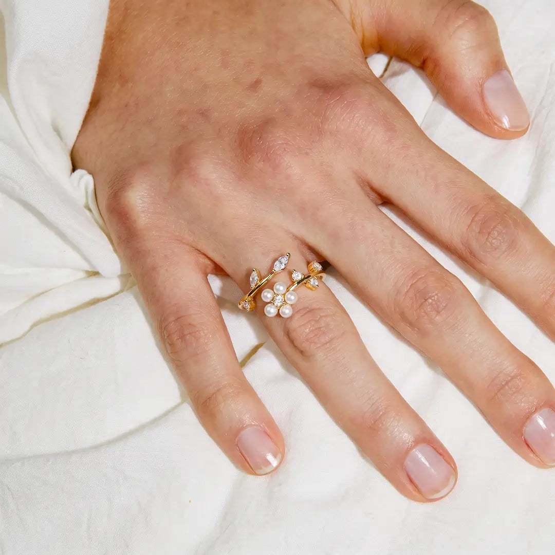 A close-up of a hand wearing a delicate gold ring adorned with small white pearls and clear crystals, resting on soft white fabric.